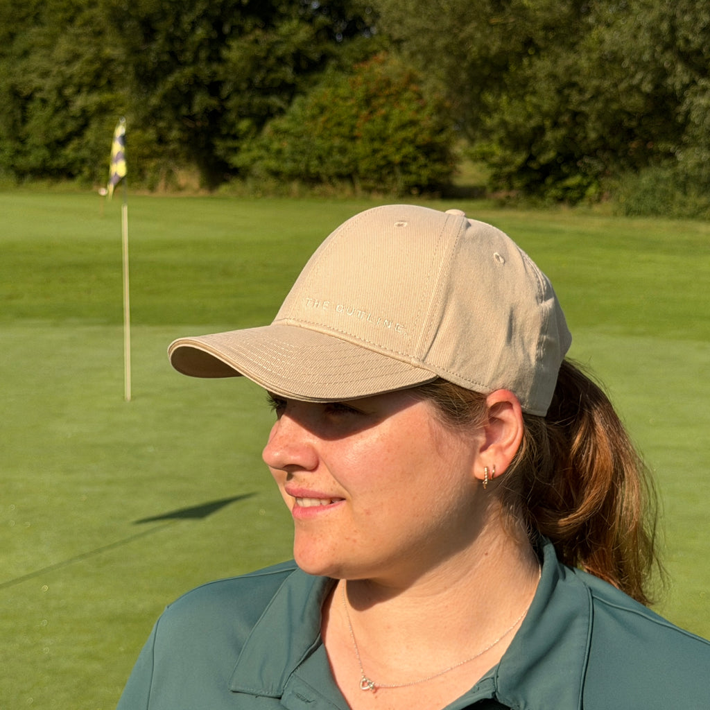 Person trägt THE CUTLINE Steady Cap in Sand mit dezentem Logo auf dem Golfplatz, Seitenansicht